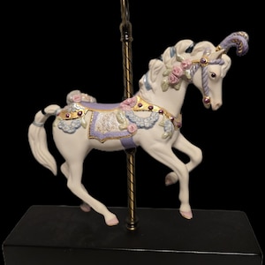 May include: A white carousel horse figurine with a gold pole and black base. The horse has a lavender saddle, pink roses, and gold accents. The horse's mane and tail are white, and it has a purple feathered headpiece.
