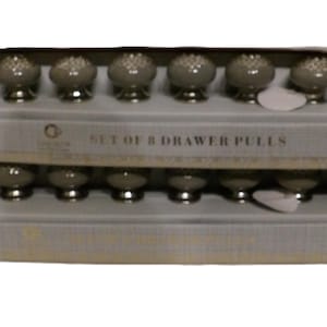 May include: A set of eight drawer pulls in a box. The drawer pulls are gray with a decorative pattern on top and a silver base. The box is light gray and has the text "SET OF 8 DRAWER PULLS".