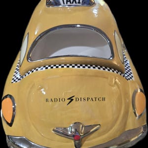 May include: A yellow ceramic taxi cab cookie jar with a glossy finish. The top features a white sign with the word TAXI. The hood displays the text RADIO DISPATCH with a lightning bolt icon. It has black and white checkered trim and silver accents.