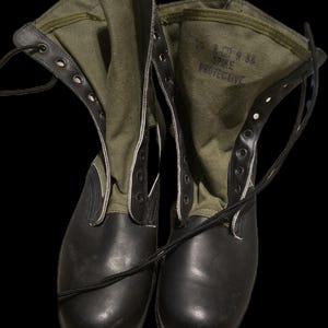 May include: A pair of black rubber boots with olive green canvas uppers and black laces. The interior of the boot features the printed text 10 R 9 88 SPIKE PROTECTIVE. These tall boots have a smooth black toe cap and a textured black rubber sole.