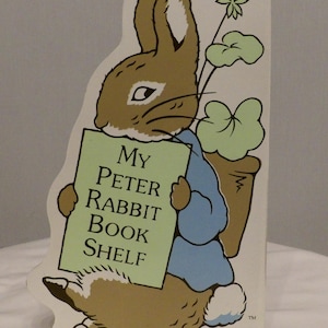 May include: A Peter Rabbit book shelf featuring a cartoon illustration of Peter Rabbit holding a sign that reads "My Peter Rabbit Book Shelf". The rabbit is brown with a blue jacket and is carrying a pot of green plants.