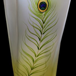May include: A tall, frosted glass vase with a hand-painted peacock feather design. The feather is depicted in shades of green, yellow, blue, and black. The vase has a rectangular shape with slightly flared sides, and the background is black.