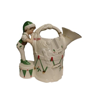 Könnte beinhalten: Weißer Keramik-Weihnachtskrug mit einer dekorativen Elfenfigur. Der Krug ist wie eine Trommel und ein Musikinstrument geformt, mit roten, grünen und goldenen Akzenten. Der Elf trägt einen grünen Hut und eine grüne Weste.