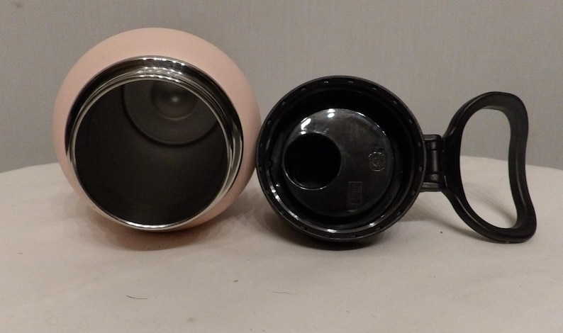 K&ouml;nnte beinhalten: Eine rosa isolierte Wasserflasche mit schwarzem Deckel und Griff. Das Innere der Flasche ist dunkel und kontrastiert mit der hellrosa Au&szlig;enseite. Der Deckel ist ge&ouml;ffnet und gibt die &Ouml;ffnung der Flasche frei.