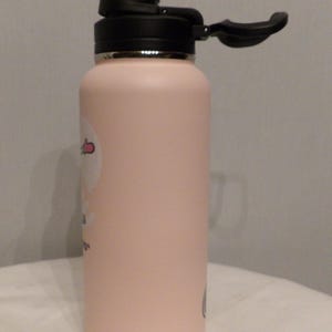 K&ouml;nnte beinhalten: Eine hellrosa isolierte Wasserflasche mit schwarzem Deckel und Griff. Die Flasche hat eine zylindrische Form und eine matte Oberfl&auml;che. Auf der Seite ist ein kleiner Aufkleber zu sehen. Der Deckel ist ge&ouml;ffnet und gibt den Trinkausguss frei.