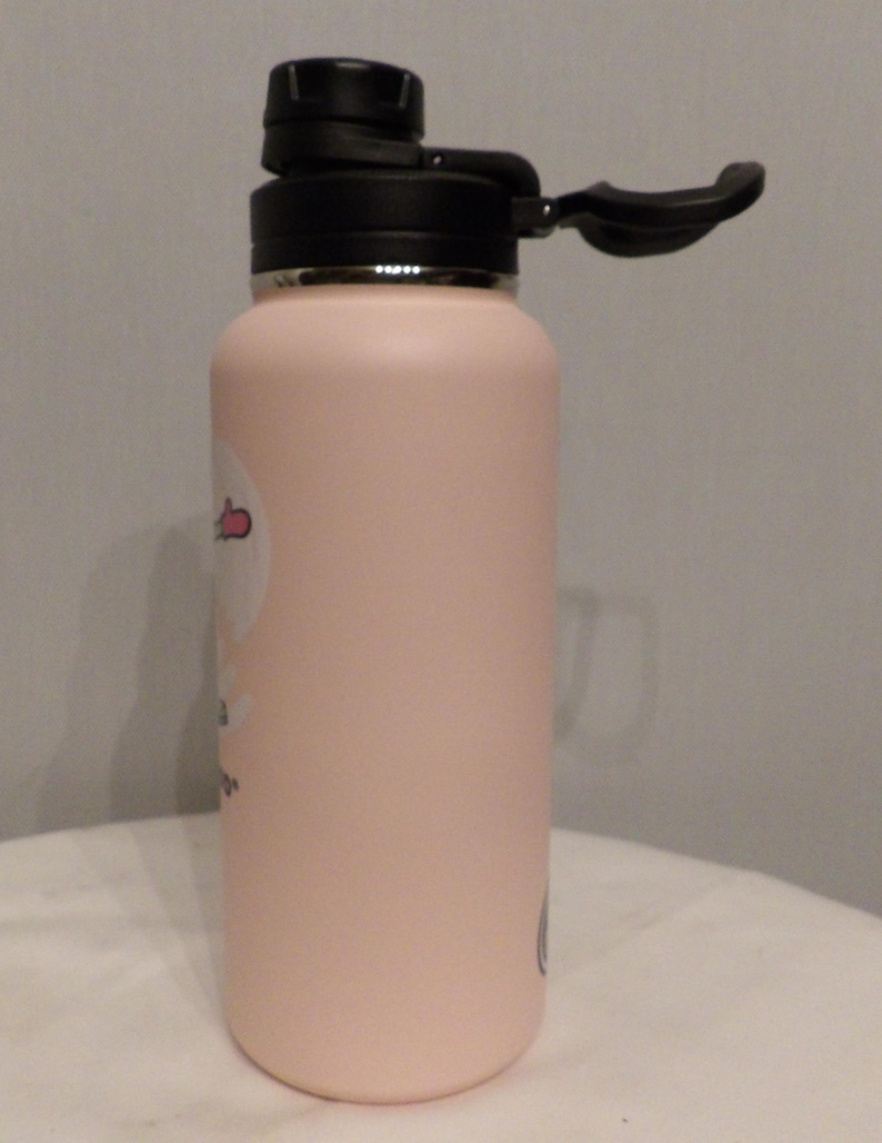 K&ouml;nnte beinhalten: Eine hellrosa isolierte Wasserflasche mit schwarzem Deckel und Griff. Die Flasche hat eine zylindrische Form und eine matte Oberfl&auml;che. Auf der Seite ist ein kleiner Aufkleber zu sehen. Der Deckel ist ge&ouml;ffnet und gibt den Trinkausguss frei.