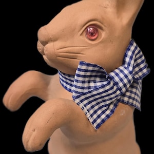 May include: A ceramic bunny figurine in a standing pose, with a light brown finish and pink eyes. It wears a blue and white gingham bow tie. The background is black.
