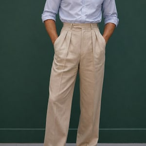 Puede incluir: Una camisa de manga larga de color azul claro con botones y cuello, junto con un pantalón beige de talle alto con pliegues. La camisa tiene un bolsillo frontal y el logotipo de Harrington Clothiers es visible arriba a la izquierda. Zapatos de cuero marrón.