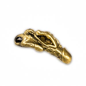 Può includere: Ciondolo color oro raffigurante una figura umana reclinata su una forma fallica. Il ciondolo ha un anello per il fissaggio a una catena o un cordino. Il design è dettagliato e strutturato.