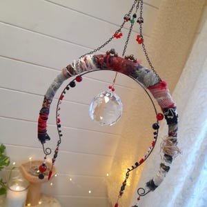 Pode incluir: Móvel artesanal com uma bola de cristal suspensa dentro de uma moldura circular. A moldura é envolta em tecido com padrões vermelhos, pretos e brancos, adornada com contas vermelhas e pretas. O móvel pende de uma corrente.