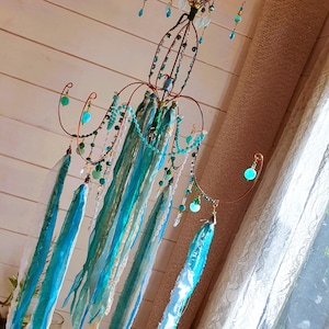 Puede incluir: Una lámpara de araña artesanal de alambre de cobre con cuentas y cintas turquesas y verdes. La lámpara de araña presenta un diseño central con brazos curvos adornados con cuentas y cintas colgantes en tonos azules y blancos. La estética general es caprichosa y decorativa.