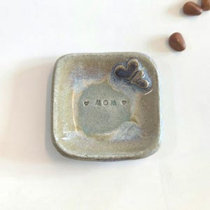 May include: Square ceramic dish with a light brown and blue glaze. The dish has the word "MOM" stamped in the center and two heart-shaped embellishments. The dish is a decorative item.