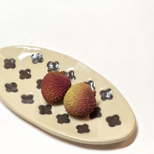 May include: An oval-shaped ceramic dish with a cream-colored glaze and a repeating pattern of dark brown, four-petaled flower motifs. Two textured, reddish-pink lychee fruits are resting in the center of the dish against a plain white background.