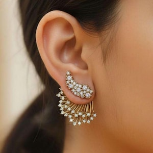 May include: A gold-toned ear cuff earring with a curved top section covered in small, clear crystals. The lower section features a fan-like design with dangling crystal accents, creating a glamorous look. The earring is designed to be worn on the ear.