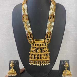 May include: Gold-toned necklace and earring set with intricate religious figures and designs. The necklace features multiple strands of white beads and a large pendant with detailed carvings. The earrings have a matching design. The set is displayed on a black mannequin.