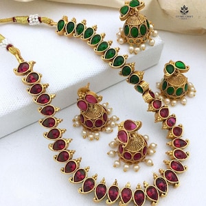 May include: A gold-toned jewelry set featuring a necklace and two pairs of earrings. The necklace and earrings are adorned with teardrop-shaped, deep red gemstones and smaller green gemstones. The earrings have a bell-like design with small white pearls.