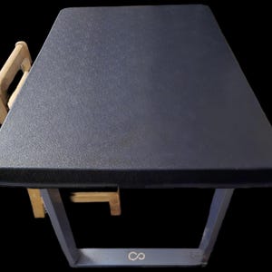 May include: A dark gray, trapezoid-shaped table with a matching chair. The table has a textured surface and a metal frame. The chair is made of light-colored wood. The table is approximately 30 inches long.