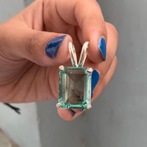 May include: A rectangular, light teal gemstone pendant with a silver bail. The gemstone is faceted, and the silver bail has a V-shape. The pendant is held up by a hand with blue nail polish.