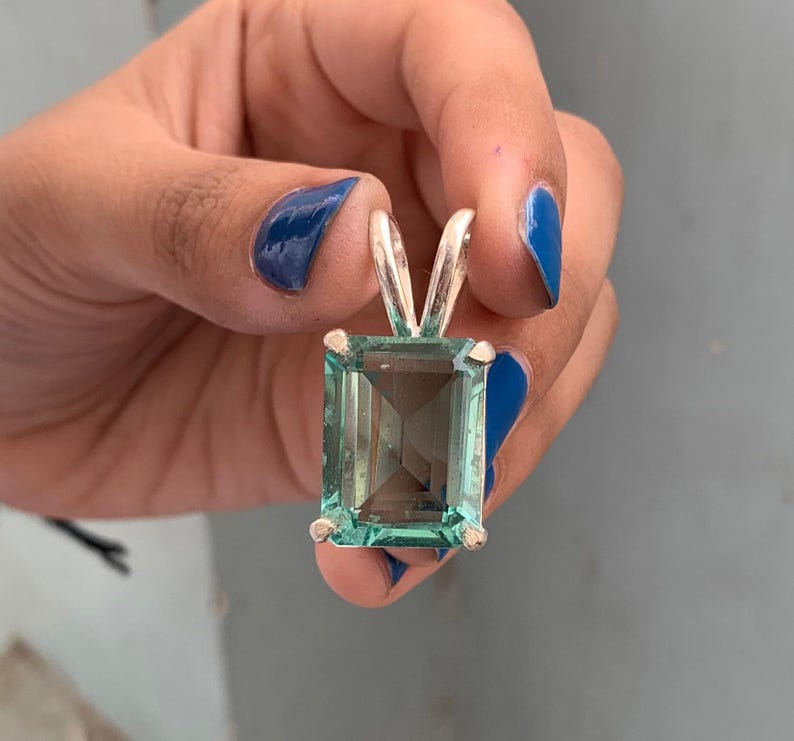 May include: A rectangular, light teal gemstone pendant with a silver bail. The gemstone is faceted, and the silver bail has a V-shape. The pendant is held up by a hand with blue nail polish.