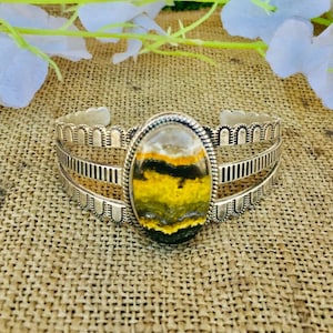 May include: Silver cuff bracelet with an oval-shaped, multi-colored stone. The stone features yellow, black, and white hues. The bracelet has a detailed design with decorative cutouts and a scalloped edge.