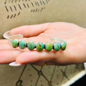 May include: Silver cuff bracelet with seven oval turquoise stones. The stones are a mix of green and blue hues, set in individual silver bezels. The bracelet has a simple, elegant design, suitable for everyday wear.