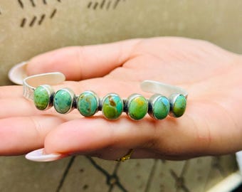 May include: Silver cuff bracelet featuring seven oval turquoise stones. The stones are a mix of green and blue hues, set in individual silver bezels. The bracelet has a simple, elegant design, perfect for everyday wear.