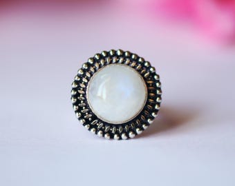 May include: A silver-toned ring with a large, round, white gemstone center. The gemstone is surrounded by a beaded border. The ring is set against a soft, blurred background with pink and white hues.