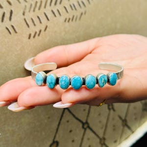 Può includere: Un bracciale a polsino in argento con sette pietre turchesi ovali. Il bracciale ha un design strutturato sulla fascia ed è mostrato su una mano. Le pietre turchesi sono di un blu vibrante. È un gioiello.