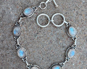 Op de afbeelding: Een zilveren armband met ovale maansteen cabochons. De stenen zijn gezet in sierlijke bezels en verbonden door kleine zilveren connectoren. De armband heeft een tuimelsluiting. De maanstenen hebben een blauw-witte iriserende gloed.