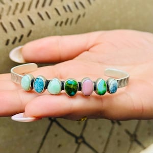 May include: A silver cuff bracelet featuring seven oval-shaped gemstones in various colors, including turquoise, pink, and green. The bracelet has a textured design on the outer edges and is a piece of jewelry.