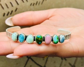 May include: A silver cuff bracelet featuring seven oval-shaped gemstones in various colors, including turquoise, pink, and green. The bracelet has a textured design on the outer edges and is a piece of jewelry.
