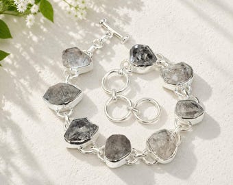May include: A silver-toned chain bracelet featuring seven irregular, raw, translucent gray stones set in metal bezels. The bracelet includes a toggle clasp with a T-bar and a circular ring, connected by polished silver-colored metal links.