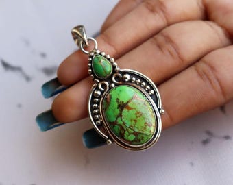 May include: A silver pendant with a large oval green stone and a smaller green stone above it. The stones have brown veining. The pendant has a decorative silver border with small beaded accents. The pendant is held in a hand.