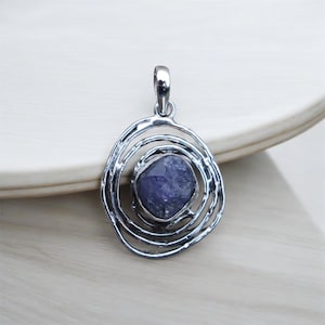 Peut inclure: Pendentif argenté avec une pierre brute bleu foncé au centre. Le pendentif présente un design ajouré circulaire avec des anneaux concentriques. La bélière est une simple boucle. Le pendentif est présenté sur une surface en bois clair.