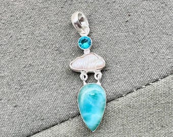 May include: A silver pendant featuring a teardrop-shaped, turquoise-colored gemstone, a white, cloud-shaped stone, and a small, round, blue gemstone. The pendant has a loop at the top for a chain.