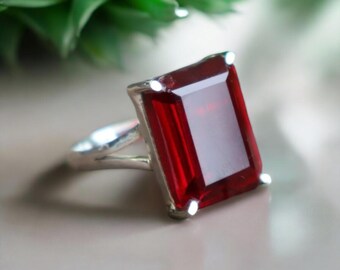 Könnte beinhalten: Ein Silberring mit einem großen, rechteckigen, tiefroten Edelstein. Der Edelstein ist in einem Silberband mit geteiltem Schaftdesign gefasst. Der Ring wird vor einem neutralen Hintergrund mit grünem Laub im Hintergrund präsentiert.