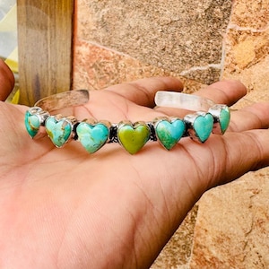 May include: Silver cuff bracelet featuring heart-shaped turquoise and green stones. The bracelet has a simple, open design with the heart-shaped stones set in individual silver bezels. The turquoise stones have a mottled appearance, while one stone is green.