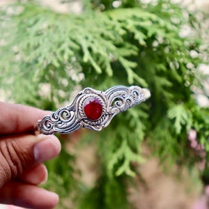 May include: Silver cuff bracelet with a central oval, deep red gemstone. The bracelet has intricate, swirling filigree patterns. The background is a blurred view of green foliage.