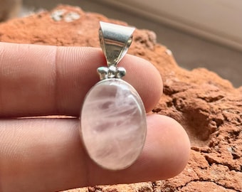 May include: Oval rose quartz pendant with a silver bail. The pendant has a soft pink hue with subtle internal patterns. The silver bail is attached to the top of the pendant. The pendant is held in a hand.