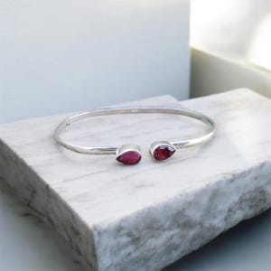 Könnte beinhalten: Ein silbernes Armreif-Armband mit zwei tropfenförmigen, tiefroten Edelsteinen. Das Armband wird auf einer weiß-grauen Marmoroberfläche präsentiert, die sein elegantes Design und die leuchtende Farbe der Steine hervorhebt.