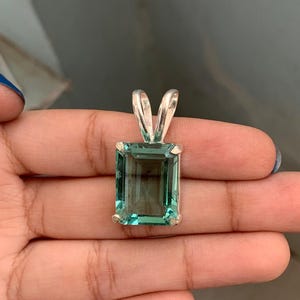 May include: A rectangular, teal-colored gemstone pendant with a silver bail. The gemstone is set in a silver frame and has a clear, transparent appearance. The pendant is held in a hand.