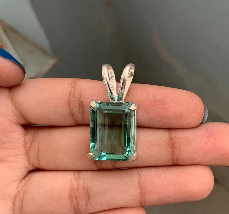 May include: A rectangular, teal-colored gemstone pendant with a silver bail. The gemstone is set in a silver frame and has a clear, transparent appearance. The pendant is held in a hand.