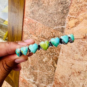 May include: A silver barrette featuring heart-shaped turquoise and green stones. The barrette has a row of six heart-shaped stones, with the center stone being green. The other stones are turquoise. The barrette is held by a hand.