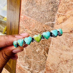 May include: A silver barrette featuring heart-shaped turquoise and green stones. The barrette has a row of six heart-shaped stones, with the center stone being green. The other stones are turquoise. The barrette is held by a hand.