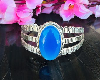 May include: Silver cuff bracelet with a large, oval, bright blue gemstone. The bracelet has a decorative design with three bands and scalloped edges. Pink flowers are in the background.