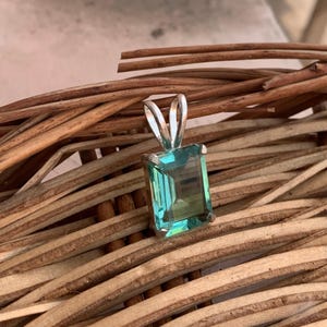 May include: A silver pendant with a rectangular, teal-colored gemstone. The pendant is set against a backdrop of woven, brown wooden sticks. The gemstone has a clear, polished appearance.