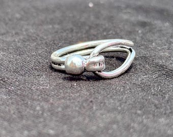 Pode incluir: Um anel prateado com um design único. O anel apresenta uma pequena conta redonda e uma banda em laço que envolve o dedo. O anel está sobre um fundo cinza escuro.