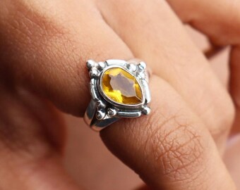 May include: A silver ring featuring a pear-shaped, golden-yellow gemstone. The gemstone is set in a decorative, square-shaped setting with small, round accents. The ring is shown on a finger, highlighting the jewelry's design and color.