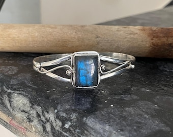 May include: A silver bracelet featuring a rectangular, iridescent blue gemstone. The bracelet has a delicate, open design with curved lines and small decorative elements. The gemstone's colour shifts with light.