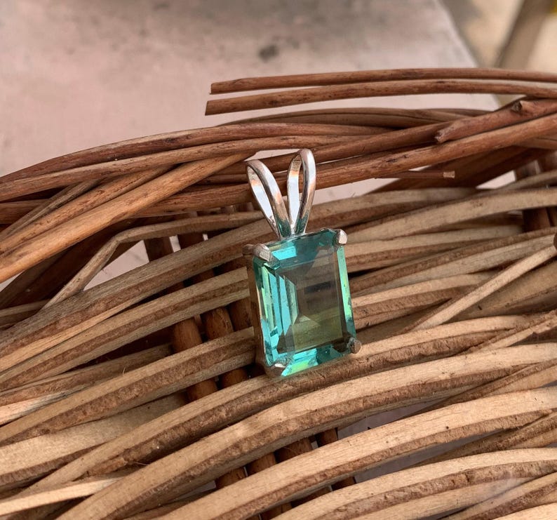 May include: A silver pendant with a rectangular, teal-colored gemstone. The pendant is set against a backdrop of woven, brown wooden sticks. The gemstone has a clear, polished appearance.